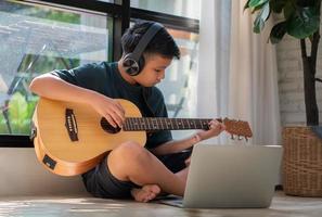 niño asiático tocando la guitarra y viendo un curso en línea en una computadora portátil mientras practica para aprender música o instrumentos musicales en línea en casa. los estudiantes varones estudian en línea con profesores de videollamadas que tocan la guitarra. foto