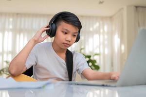 Asian boy playing guitar and watching online course on laptop while practicing for learning music or musical instrument online at home. Boy students study online with video call teachers play guitar. photo