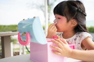 Adorable little child Asian girl paints her mouth with pink children heads and looks in the mirror. A child plays at home in a toy beauty salon. Increase learning development for preschoolers. photo