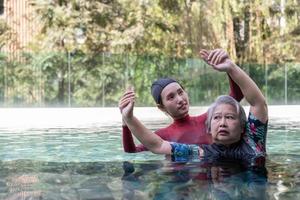 Young trainer helping senior woman in aqua aerobics and working out in the pool. old woman and mature man doing aqua aerobics exercise in swimming pool, Elderly sports, and active lifestyle concept. photo