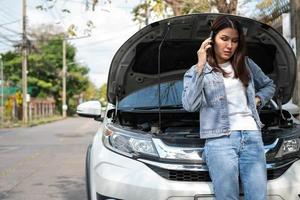 Angry Asian woman and using mobile phone calling for assistance after a car breakdown on street. Concept of vehicle engine problem or accident and emergency help from Professional mechanic photo