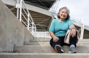 Happy and smile elderly asian woman sitting on stairs for rest after workout, jogging on morning, senior exercise outdoor for good healthy. Concept of healthcare and active lifestyle for healthy photo