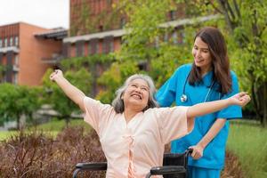 Asian careful caregiver or nurse and the happy patient in a wheelchair are walking in the garden. Concept of happy retirement with care from a caregiver and Savings and senior health insurance. photo