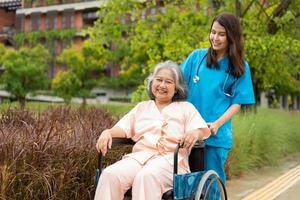 Asian careful caregiver or nurse and the happy patient in a wheelchair are walking in the garden. Concept of happy retirement with care from a caregiver and Savings and senior health insurance. photo