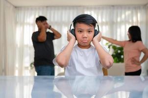 Asian boy kid wearing headphones and play loud music. so as not to hear quarrel while parents having fight or quarrel conflict at home. Unhappy problem in family, Domestic problems in the family. photo