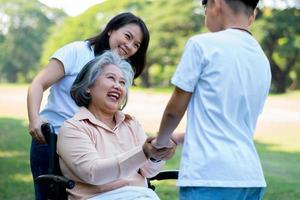 Happy senior Asian grandmother uses wheelchair with her daughter and grandchild in park, Grandson came to visit elderly grandmother and hold hand. Concept of happy family, good relationship together photo