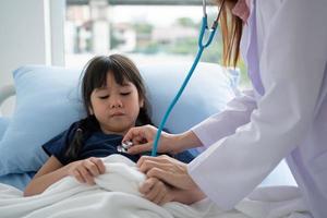 Asian woman pediatrician doctor hold stethoscope for exam a little girl patient and heck heart lungs of kid, Good family doctor visiting child at home, Healthcare and medicine for childhood concept. photo