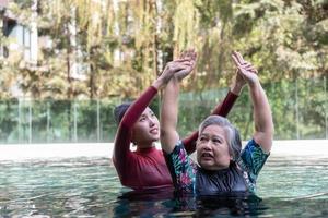 Young trainer helping senior woman in aqua aerobics and working out in the pool. old woman and mature man doing aqua aerobics exercise in swimming pool, Elderly sports, and active lifestyle concept. photo