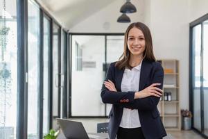 un elegante blanco mujer de negocios en un negro traje en pie dentro el oficina en manos con un móvil teléfono, tableta, y ordenador portátil esperando para clientes a contacto, negocio personas concepto. foto