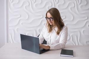 A young business woman with glasses is sitting at a white desk and looking at a laptop photo