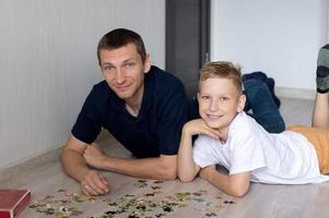 A cute boy with a cheerful dad is collecting a puzzle lying on the floor in a room photo