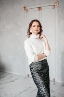 A girl in a white sweater and a shiny skirt stands in the interior and looks at the camera photo