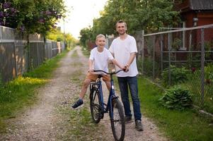 A cute boy in a white T-shirt learns to ride a bike with his dad and laughs. Dad teaches his son.