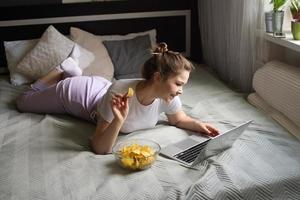 A cute girl is lying on the bed eating chips and looking at a laptop photo