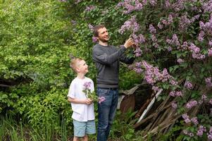 papá soportes cerca un floreciente lila arbusto y ayuda su hijo a recoger un ramo de flores foto