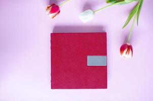 un rojo cuero libro con un en relieve sitio para texto, mentiras en un púrpura antecedentes siguiente a flores y tulipanes foto