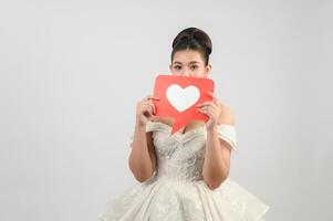 hermosa novia asiática sonriendo y posando con el signo del corazón en el fondo blanco foto