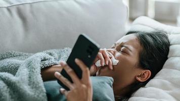 Asian woman in cloak suffering from fever and flu on sofa while using mobile phone photo