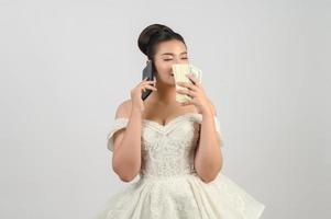 joven novia asiática hermosa que publica con un teléfono inteligente y un billete en la mano foto