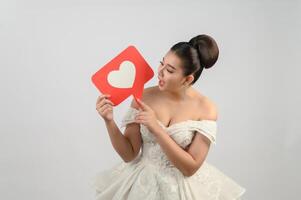 hermosa novia asiática sonriendo y posando con el signo del corazón en el fondo blanco foto