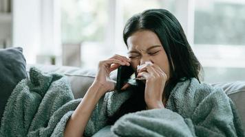 Asian woman in cloak suffering from fever and flu on sofa while talking with mobile phone