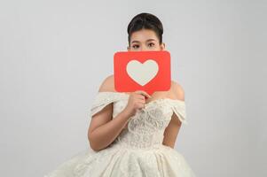 hermosa novia asiática sonriendo y posando con el signo del corazón en el fondo blanco foto