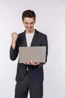 Portrait of young smiling businessman holding laptop in hands, typing and browsing web pages while doing a winning closed fist gesture isolated on white background photo
