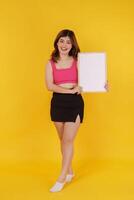 Portrait of Happy young woman holding an empty white placard over isolated yellow background. photo