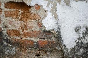 Old cement wall cracked to reveal red bricks inside, poor quality construction. photo