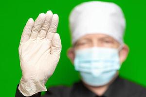 Doctor male in surgical face mask on defocus foreground put forward palm of his hand in protective latex glove foreground, waves by his hand, hello or stop sign photo