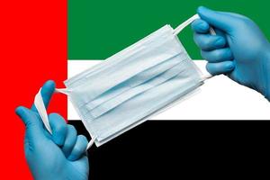 Medic holding respiratory face mask in hands in blue gloves on background national flag of United Arab Emirates UAE. Concept coronavirus quarantine photo