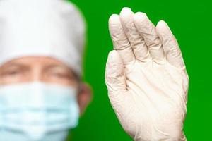 Male doctor in surgery face mask on defocus foreground put forward palm of his hand in medical protective glove foreground, waves his hand, stop or hello sign photo