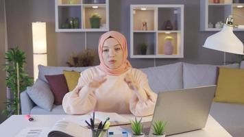 giovane ragazza nel hijab è studiando a casa e è annoiato. musulmano alunno guardare a il computer portatile e libri è annoiato con studiando. video