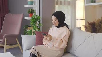 Young Arab Muslim woman in hijab uses smartphone at home. Woman in headscarf is texting on the phone and smiling. video