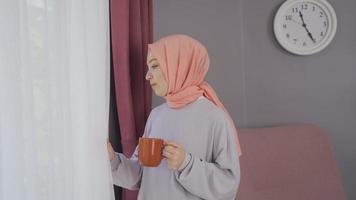 musulman femme regards en dehors le la fenêtre. Jeune femme dans une foulard est en train de regarder en dehors le fenêtre à maison. video