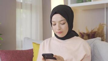 kvinna i slöja är textning på de telefon och leende. ung muslim kvinna textning på telefon på Hem. video