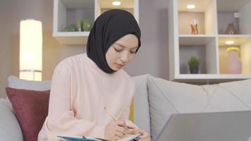árabe negocio mujer vistiendo un Pañuelo es trabajando desde hogar. joven hijab mujer mirando a ordenador portátil a hogar y escritura alguna cosa en papel. video