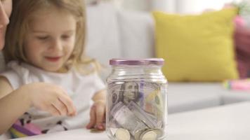 Little girl puts money in piggy bank with her mother. Her mother teaches her daughter how to save money. Caring parent teaches kid to save money, think about future, manage personal finance. video