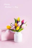 un ramo de flores de vistoso tulipanes en un moderno blanco florero en un rosado antecedentes con un regalo caja. el texto de un contento de la madre día. vertical vista. foto