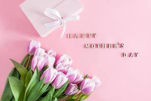 festive background with lilac tulips and a gift box with a bow. text in wooden letters of Happy Mother's Day. top view. pink background. photo