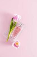 a bottle of delicate women's perfume or spray lies on a glass relief tray with a tulip. vertical view. pink background. aroma presentation.