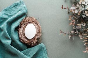 White chicken egg in a nest on a linen napkin and a branch of eucalyptus on a green background. Eco friendly happy easter. Top view. photo