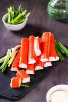 Spicy crab sticks and green onions on a stone board and sauce in a bowl on the table. Vertical view photo