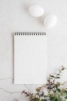 Notepad for writing, white eggs and eucalyptus branch on a light textured background. Happy eco easter invitation. Top and vertical view.