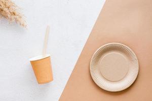 The concept of eco-friendly utensils for food. Cardboard cup, drinking straw, plate and ear of grass on plaster and beige background. Top view. photo