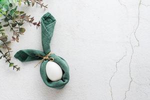 A white chicken egg is wrapped in a napkin in the form of a rabbit and a branch of eucalyptus on a light texture background. Eco friendly happy easter. Top view. photo