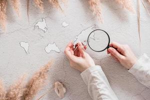 Flat lay concept travel. Female hands hold templates of continents and a magnifier surrounded by grass on a textured concrete background. photo