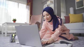 hembra musulmán estudiante estudiando utilizando ordenador portátil y libros. un musulmán estudiante sentado en el piso a hogar es estudiando para su lecciones video