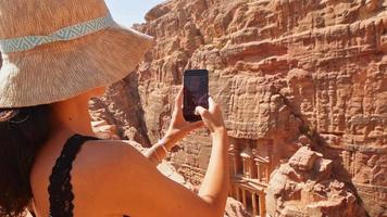 Gorgeous caucasian woman tourist sit on viewpoint in Petra ancient city over Treasury or Al-khazneh take smartphone photo. Jordan, one of seven wonders. UNESCO World Heritage site. video