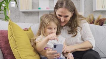 madre y hija mirando a el teléfono a hogar. madre demostración su pequeño hija alguna cosa en el teléfono. video